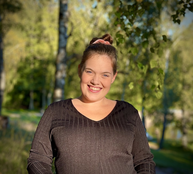 Leende person utomhus i solljus med gröna träd i bakgrunden