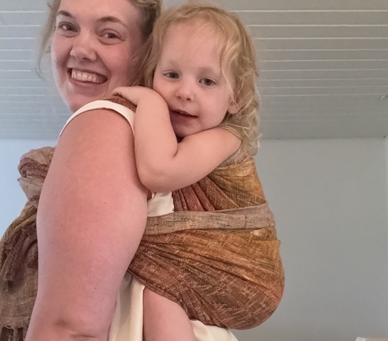 En leende vuxen bär ett litet barn i en brun bärsjal inomhus